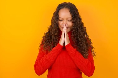 Kapalı alanda, kırmızı kazak giyen, sarı arka planda duran, yoga ve meditasyon yapan, namaste 'de avuç içlerini bir arada tutan sakin, rahat ve huzurlu görünen bir kadın..