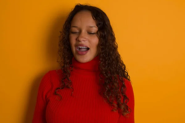 Stressful  woman wearing red sweater standing against yellow background screams in panic, closes eyes in terror, keeps hands on head, finds out terrified news, can\'t believe it.