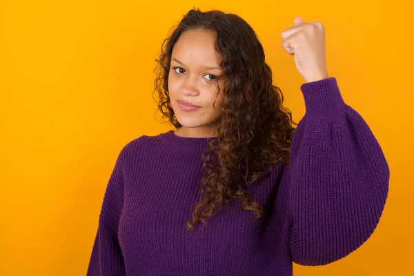 Fierce  woman wearing purple sweater standing against yellow background holding fist in front as if is ready for fight or challenge, screaming and having aggressive expression on face.