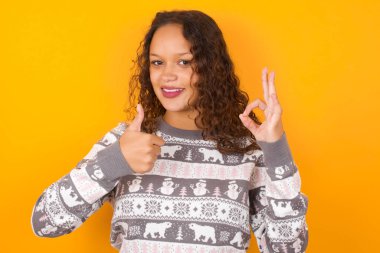  Kadın Noel süveteri giyiyor sarı stüdyo arka planında gülümsüyor ve mutlu görünüyor, gamsız ve pozitif, bir eliyle zafer ya da barış işareti yapıyor.