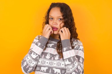 Noel süveteri giyen suskun bir kadın sarı stüdyo arka planında elleri açık, ağzı açık, şok edici haberlere tepki veriyor kameraya hayret verici bakışlar atıyor.