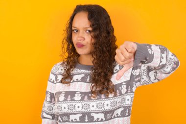  Noel süveteri giyen kadın, sarı stüdyo arka planında mutsuz, kızgın, reddedilmiş ve olumsuz davranışlar sergiliyor. Kötü ifade.