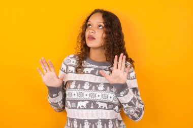  Noel süveteri giyen kadın sarı stüdyo arka planı avuçlarını ileri doğru tutar ve tavandan korkuyla bakar kendini görünmez tehlikeden korumaya çalışır. Ağzını açar..