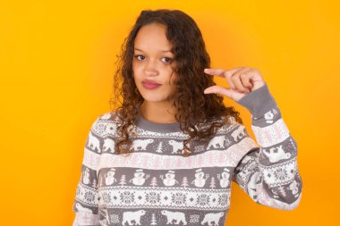  Noel süveteri giyen, sarı stüdyo arka planı olan, dudak ve el hareketleriyle hareket eden bir kadın çok küçük bir şey gösteriyor..