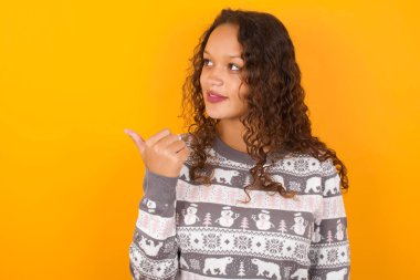  Noel süveteri giyen kadın, sarı stüdyo arka planına işaret ediyor ve reklam sunuyor.