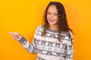 Noel süveteri giyen kadın, sarı stüdyo arka planı gülerek boş alandaki mutluluğu gösteriyor. İyi haber ya da beklenmedik satıştan etkilenmiş, indirimli alışveriş teklifini gösteriyor..