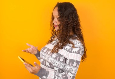 Noel süveteri giyen mutlu bir kadın, sarı stüdyo geçmişi olan, cep telefonu kullanan, cep telefonu kullanan ve cep telefonuyla mesajlaşma ya da video görüşmeleri yapan biri.