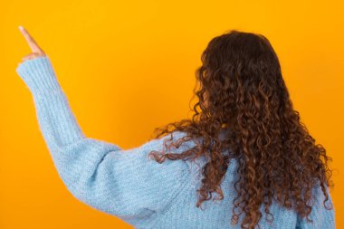 Sarı stüdyo arka planında mavi kazak giyen kıvırcık kadın fotokopi alanındaki objeyi işaret ediyor. Arkanı dön.