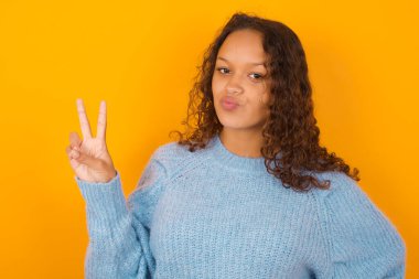   Sarı stüdyo arka planında mavi kazak giyen kıvırcık kadın barış jesti yapıyor dudaklarını katlanmış V işaretini koruyor. Vücut dili kavramı
