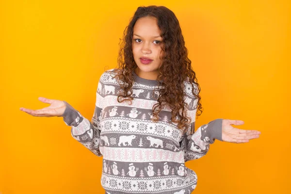 So what? Portrait of arrogant  woman wearing Christmas sweater agaist yellow studio background shrugging hands sideways smiling gasping indifferent, telling something obvious.