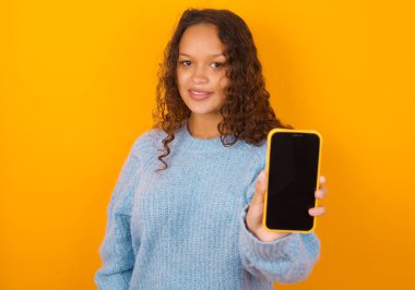 Sarı arka planda mavi süveter giyen sevimli kıvırcık kadın elinde modern bir aletle siyah ekran akıllı telefon gösteriyor.