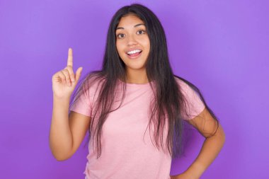 Pembe tişörtlü esmer kız mor arka plan üzerinde. Parmağını kaldırıp poz veriyor.