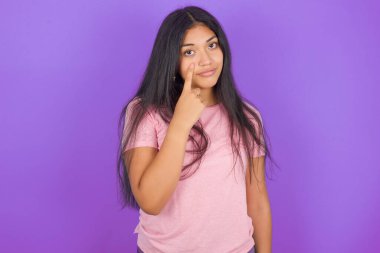 Mor arka planda pembe tişört giyen esmer İspanyol kız jestini ve şüpheli ifadesini izlediğini gösteriyor..