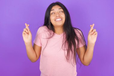 Mor arka planda pembe tişört giyen esmer İspanyol kız parmaklarını çapraz yapıp umutla gülümsüyor ve gözlerini kapıyor. Şans ve batıl inanç..