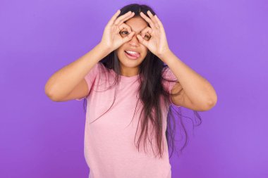 Mor arka planda pembe tişört giyen esmer İspanyol kız dürbünün dili dışarı çıkarması, gözlerin parmaklarından içeri bakması gibi güzel hareketler yapıyor. Çılgın ifade.