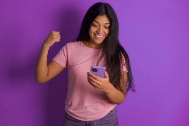 Mor arka planda pembe tişört giyen İspanyol esmer kız elinde akıllı telefonuyla iyi haberleri okuduktan sonra heyecandan yumruğunu kaldırıyor..