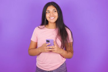 Mor arka planda pembe tişört giyen esmer İspanyol kız elinde telefonla iyi haberler okuyor ve boş alana bakıyor.