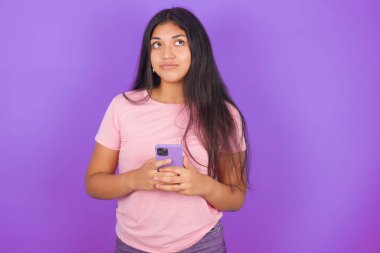 Mor arka planda pembe tişört giyen esmer İspanyol kız telefonu elinde tutuyor, iyi haberler okuyor ve boş alana bakıyor.