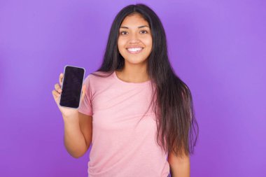 Gülümseyen esmer, pembe tişörtlü, mor arka planda esmer kız fotokopi çektir. Boş ekran ile cep telefonunu tutar