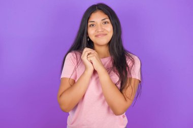 Mor arka plan üzerine pembe tişört giyen çekici İspanyol kız kameraya nazikçe gülümsüyor.