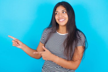 İyimser genç İspanyol kız çizgili tişört giyiyor mavi arka plan noktaları ve iki eliyle boş yere bakıyor..