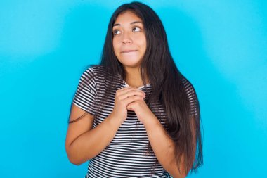 Mavi arka planda çizgili tişört giyen meraklı genç İspanyol kız ellerini çenesinin altında tutuyor, dudaklarını ısırıyor ve ilgiyle bakıyor..