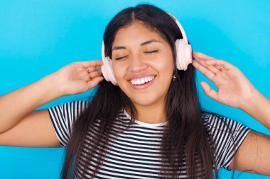 Mavi arka planda çizgili t-shirt giyen genç İspanyol kız gülümsüyor. Kablosuz kulaklıkla en sevdiği müziği dinlediği için çok mutlu. Gözlerini kapatıyor..