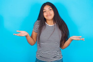 Dikkatsiz, çekici, İspanyol bir kız. Mavi arka plan üzerinde çizgili tişört giyiyor, omuzlarını silkiyor.