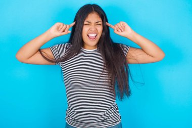 Mavi arka planda çizgili tişört giyen çılgın İspanyol kızın fotoğrafı. Çığlık atıyor ve parmakları saçları kapalı gözleri işaret ediyor.