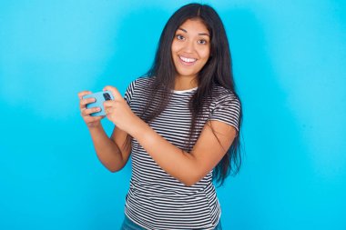 İyi bağımlı, neşeli İspanyol kız mavi arka plan üzerinde çizgili tişört giyiyor akıllı telefon kullanıyor, ağ oyunu oynuyor.