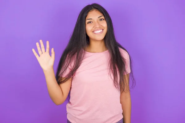 ハロー 幸せで笑顔 フレンドリーな歓迎のジェスチャーを振って紫色の背景の上にピンクのTシャツを着ているヒスパニックなブルネットガール — ストック写真