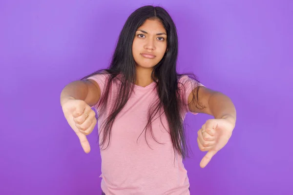 紫の背景にピンクのTシャツを着たヒスパニック系のブルネットの女の子は 2つの手で親指を下に示す動揺しています コンセプトが嫌い — ストック写真