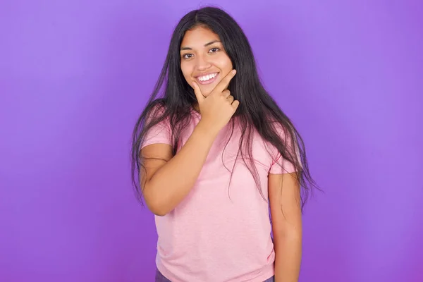 紫の背景にピンクのTシャツを着たヒスパニック系のブルネットの女の子は カメラが交差した腕と手であごに上げ笑顔で自信を持って見ています 肯定的な考え — ストック写真