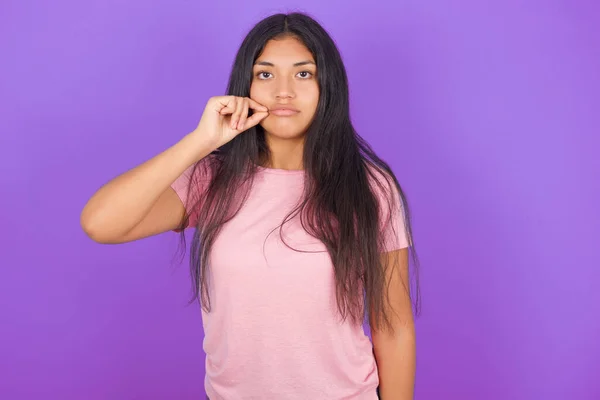 ピンクのTシャツを着たヒスパニック系のブルネットの女の子紫色の背景の口と唇の上に指でZipとしてシャットダウンします 秘密裏に黙ってタブーを語る — ストック写真