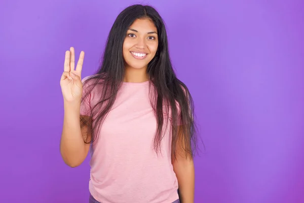 ヒスパニックブルネット女の子身に着けていますピンクTシャツ以上紫の背景ショーと指摘指番号3ながら笑顔自信と幸せ — ストック写真