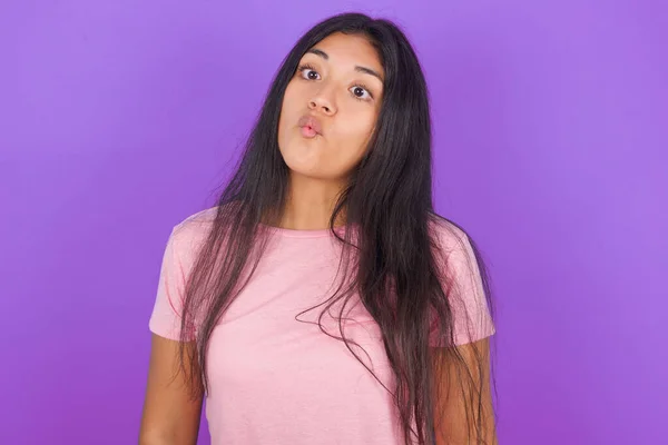 紫の背景にピンクのTシャツを着たヒスパニック系のブルネットの女の子は 狂気とコミカルなジェスチャーで魚の顔を作る 面白い表現 — ストック写真