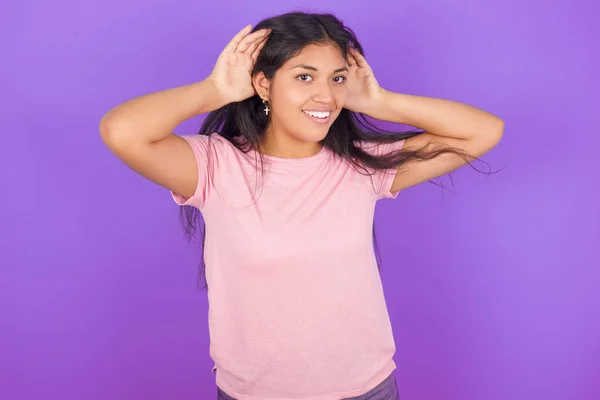 耳のジェスチャーで両手を聞こうとしている紫色の背景にピンクのTシャツを着ているヒスパニックなブルネットガールは ゴシップに興味があります アメリカ合衆国国防総省 — ストック写真