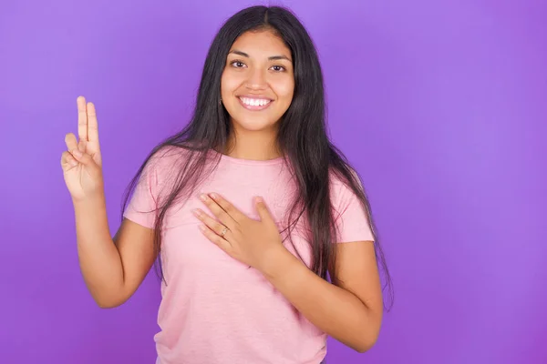 ヒスパニック系ブルネットの女の子身に着けていますピンクTシャツ以上紫の背景笑顔身に着けています手で胸と指アップ 忠誠の誓いを作ります — ストック写真