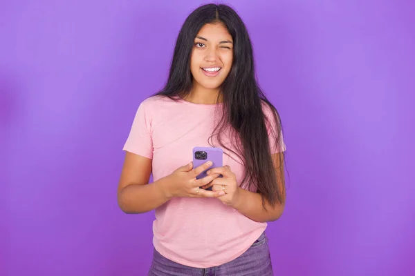 Латиноамериканская Брюнетка Розовой Футболке Фиолетовом Фоне Помощью Телефона Подмигивая Камеру — стоковое фото