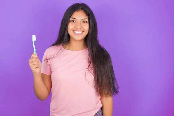 パープルの背景にピンクのTシャツを着たヒスパニック系のブルネットの女の子が歯ブラシと笑顔を持っています 歯科医療の概念 — ストック写真