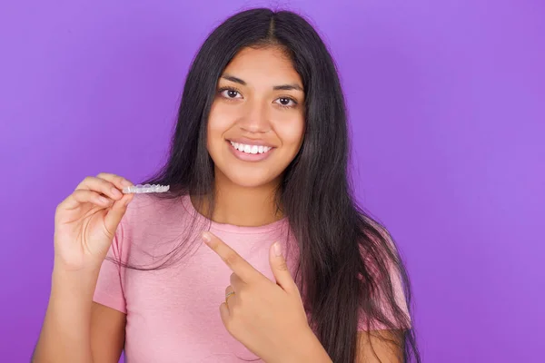 紫色の背景にピンクのTシャツを着たヒスパニック系のブルネットの女の子は 目に見えないワニを保持し それを指しています 歯科医療と信頼の概念 — ストック写真