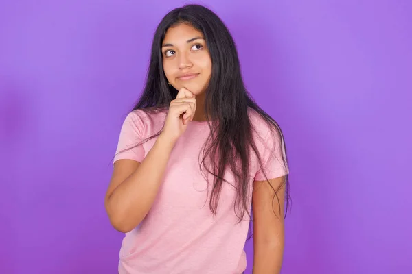 思考ヒスパニック系ブルネット女の子身に着けていますピンクTシャツ以上紫の背景保持あごと離れて見ますPensive作る素晴らしい計画 — ストック写真