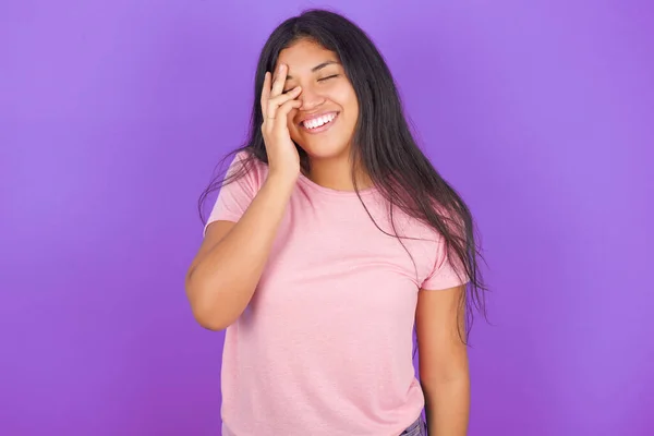 紫色の背景にピンクのTシャツを着用したヒスパニックなブルネットガールは 顔のパームと笑顔を広くし 陽性に面白いジョークポーズを聞きます — ストック写真
