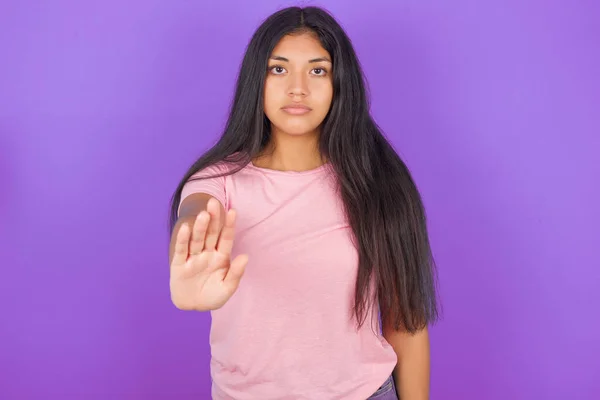 年轻的惊慌失措的女孩穿着紫色背景的粉色T恤 展示了禁止牌的标志 手心紧握着相机 表情严肃 — 图库照片