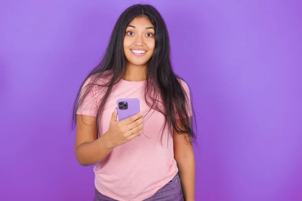 Молодая Латиноамериканка Розовой Футболке Фиолетовом Фоне Держит Руках Мобильный Телефон — стоковое фото
