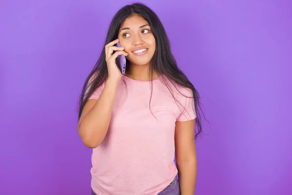 紫色の背景にピンクのTシャツを着ている幸せな幸せな若いヒスパニックの少女は素晴らしい電話会話を持っており 脇に見えます 電話で話しながら良い気分と笑顔を持っています — ストック写真