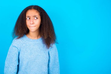 Şaşırmış afro saçlı, mavi arka planda mavi süveter giyen, dudaklarını kıvıran endişeli bakışlı, önünde korkunç bir şey gören afro-amerikalı genç kız..