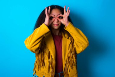 Afro saçlı, mavi arka planda sarı ceketli, neşeli Afro-Amerikan genç kız. Tamam işareti iki gözünün üstünde, gözlük takıyormuş gibi yapıyor..