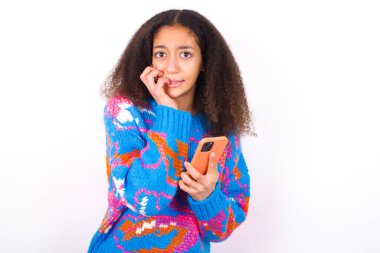 Afro saçlı, beyaz arka plan üzerine renkli süveter giyen, telefon tutan ve tırnaklarını yiyen komik Afrikalı Amerikalı kız.
