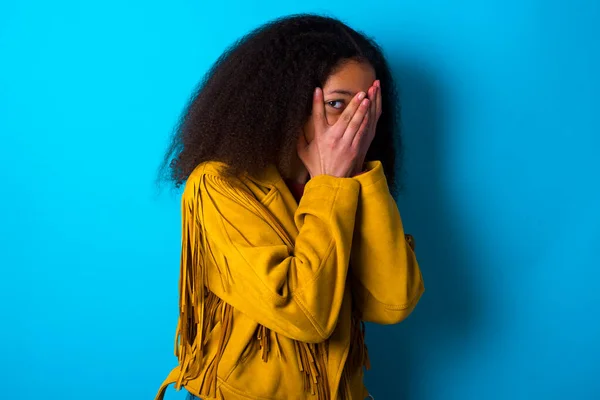 非洲裔美国少女 留着非洲式的头发 身穿黄色夹克 蓝色背景 双手捂住脸 用一只手指头朝外张望 被什么东西或某个人吓坏了 — 图库照片